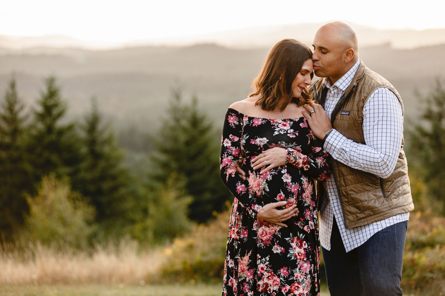 A couple embraces in a romantic mountain setting at sunset, with pine trees and rolling hills in the background.