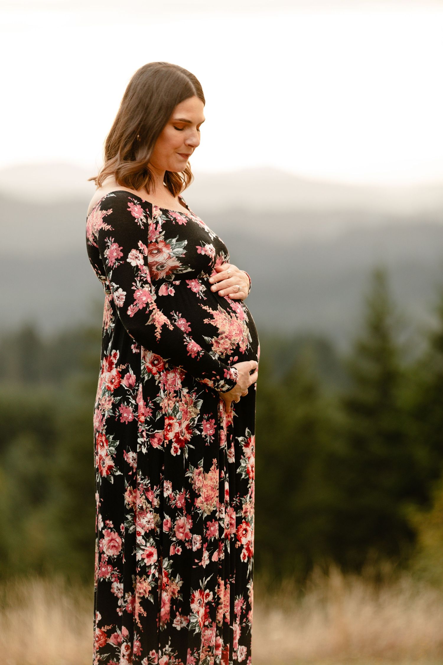 Person in black floral maxi dress poses in beautiful mountain landscape during maternity photoshoot.