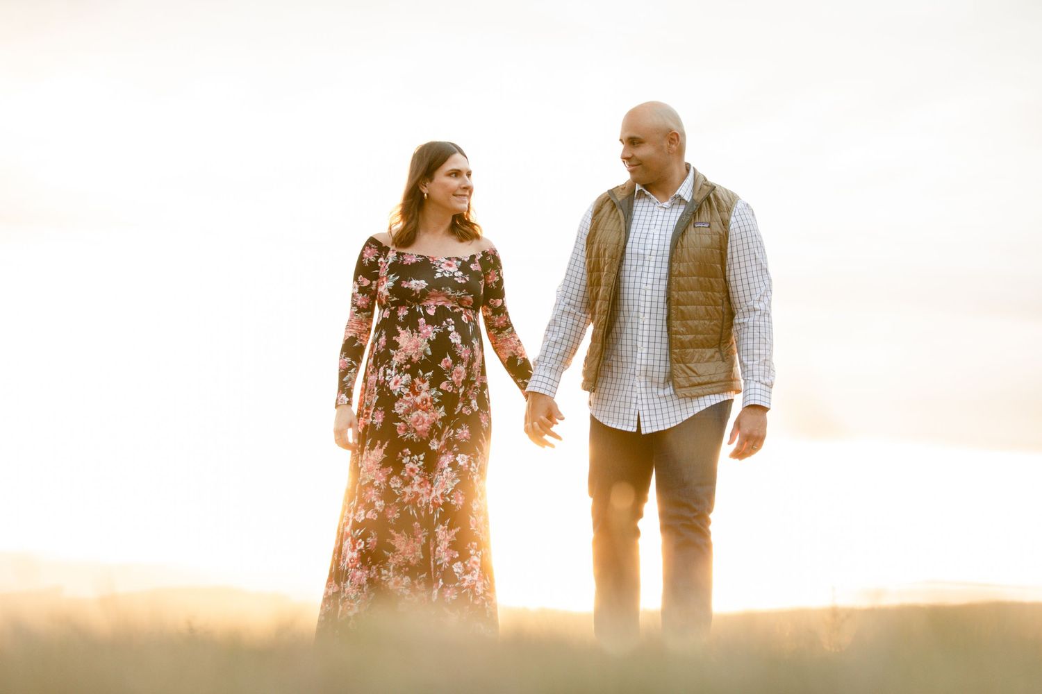 A couple holds hands during sunset with a warm backlit glow, the woman wearing a floral dress and man in casual attire.