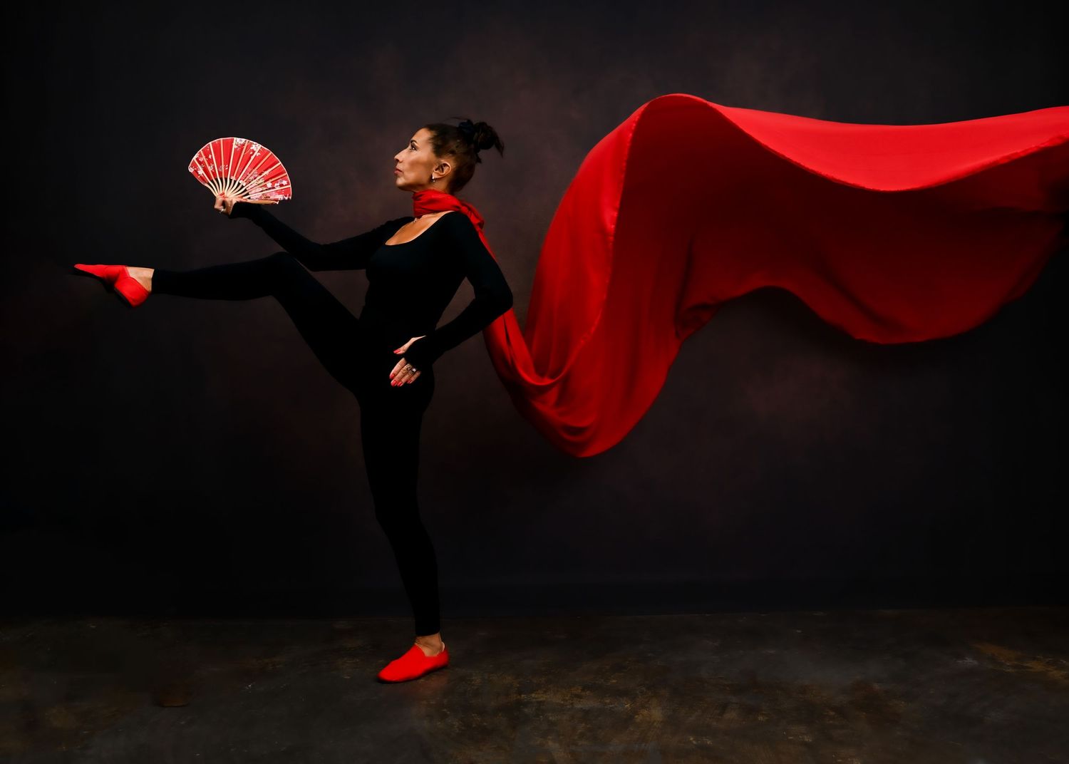 A dancer performs in a black outfit with a flowing red cape and fan against a dark background.