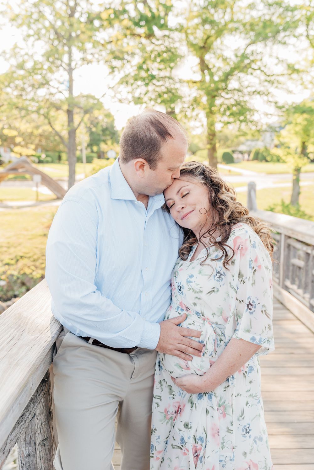 Maternity session with expecting mother standing near a serene lake and bridge in Monmouth County, NJ while her husband kisses her forehead.