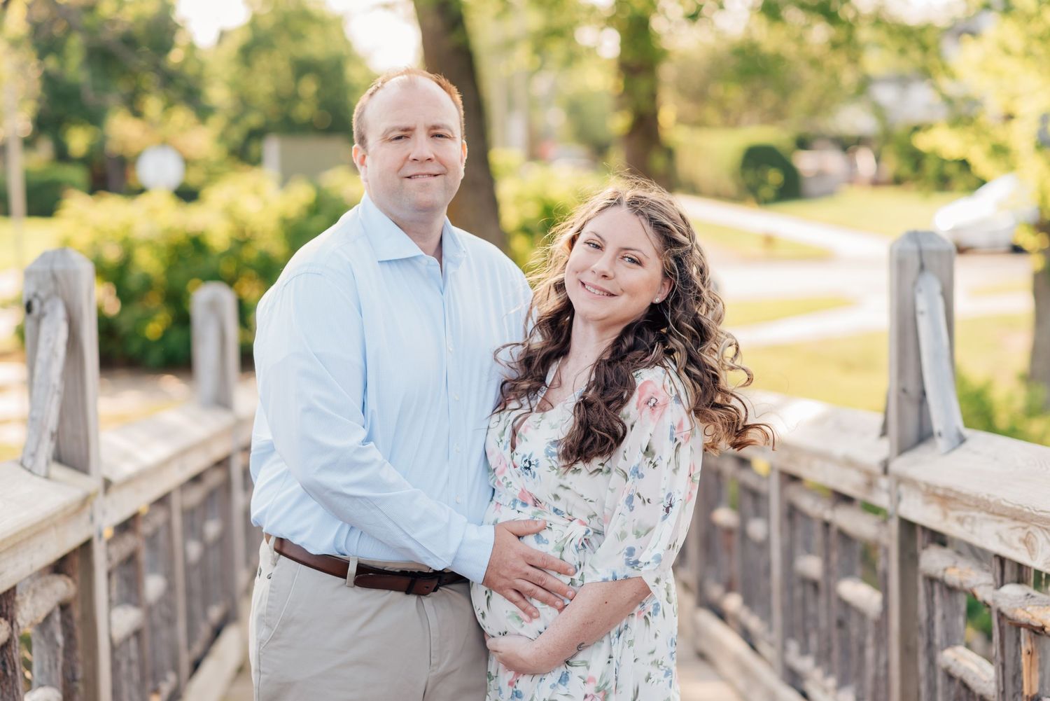 Expecting mother standing on a bridge near a lake in Monmouth County, NJ, with her husband wearing a flowing white floral gown during sunset