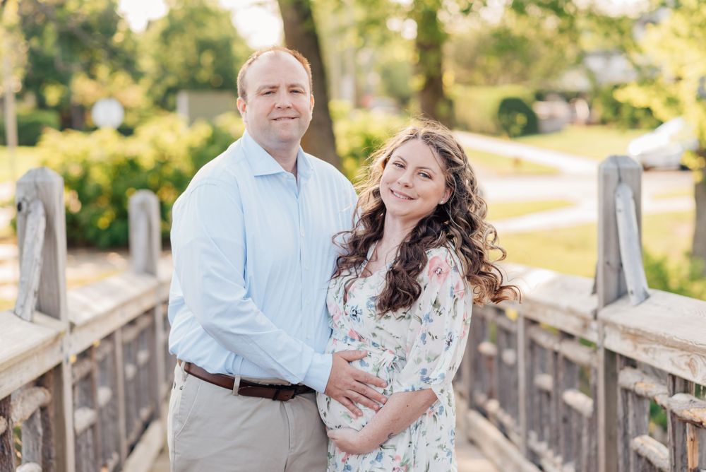 Expecting mother standing on a bridge near a lake in Monmouth County, NJ, with her husband wearing a flowing white floral gown during sunset