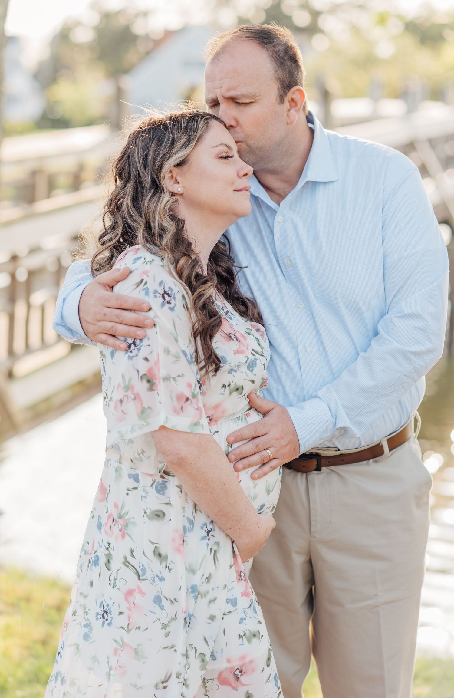 A man kisses his pregnant wife while embracing her in front of a lake in a spring lake park.