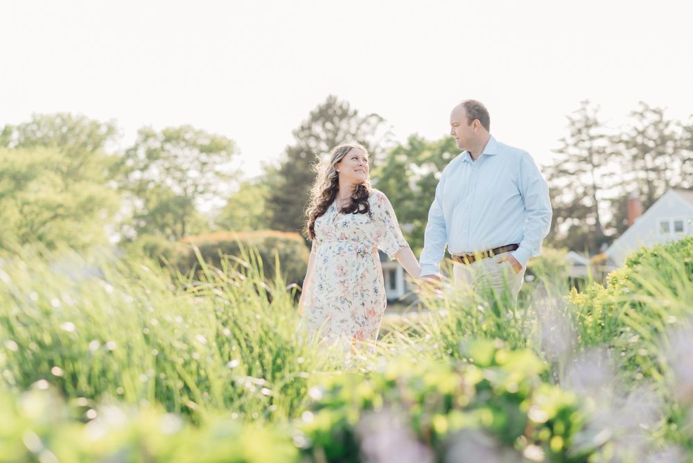 A man and pregnant women hold hands and walk along a lake with lots of flowers.