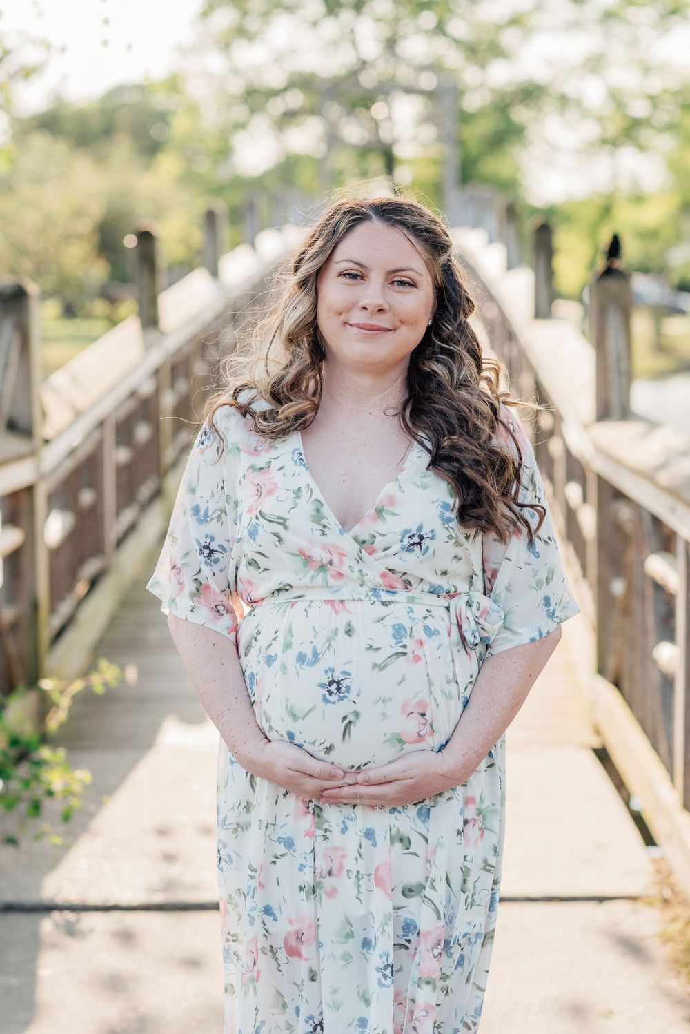 Expecting mother holds her belly in front of a bridge on a lake at Divine park in Spring Lake.