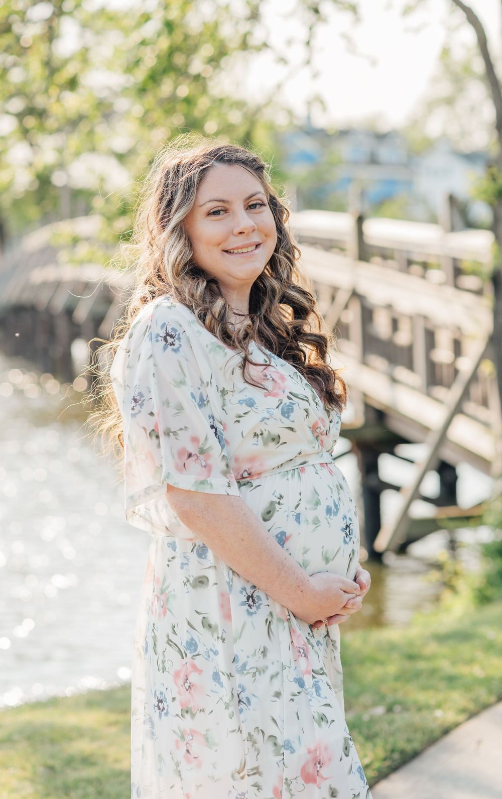 A pregnant woman holds her belly smiling in front of a bridge crossing over a lake.