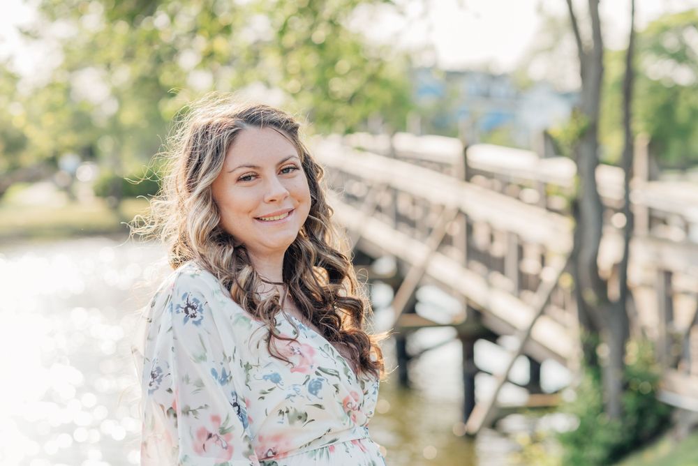 A portrait of a woman in a flower dress smiles in front of a bridge and lake.