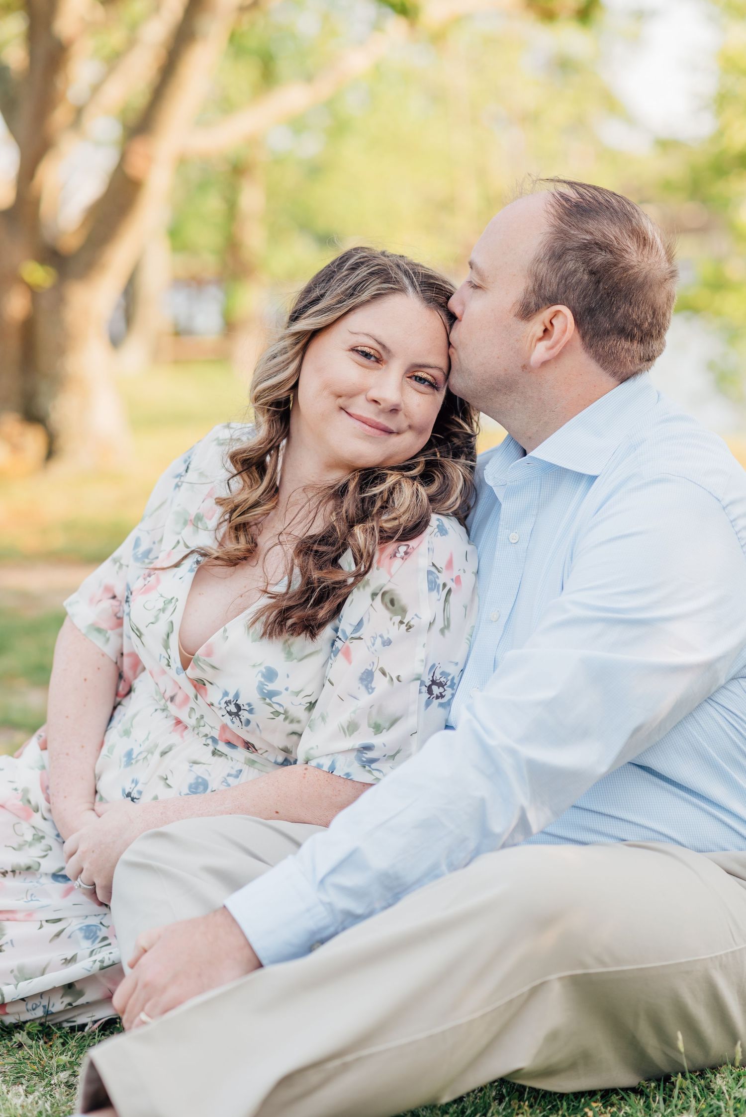 A man kisses a pregnant woman while sitting on the grass in a park.
