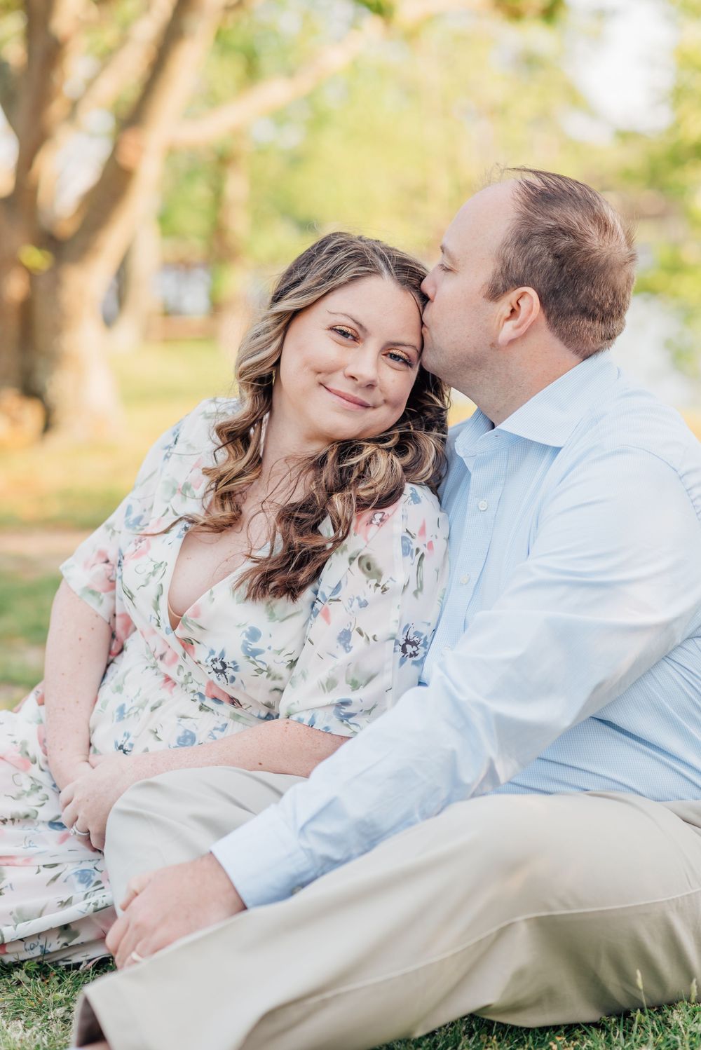 A man kisses a pregnant woman while sitting on the grass in a park.
