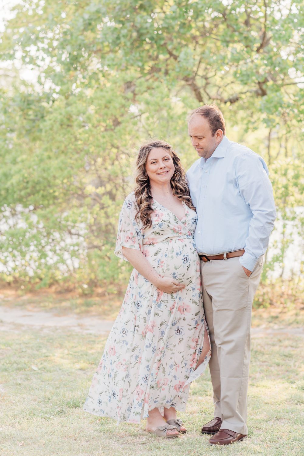 Portrait of a man and a pregnant woman outdoors leaning into each other looking down with her hands placed on her stomach and he is smiling.