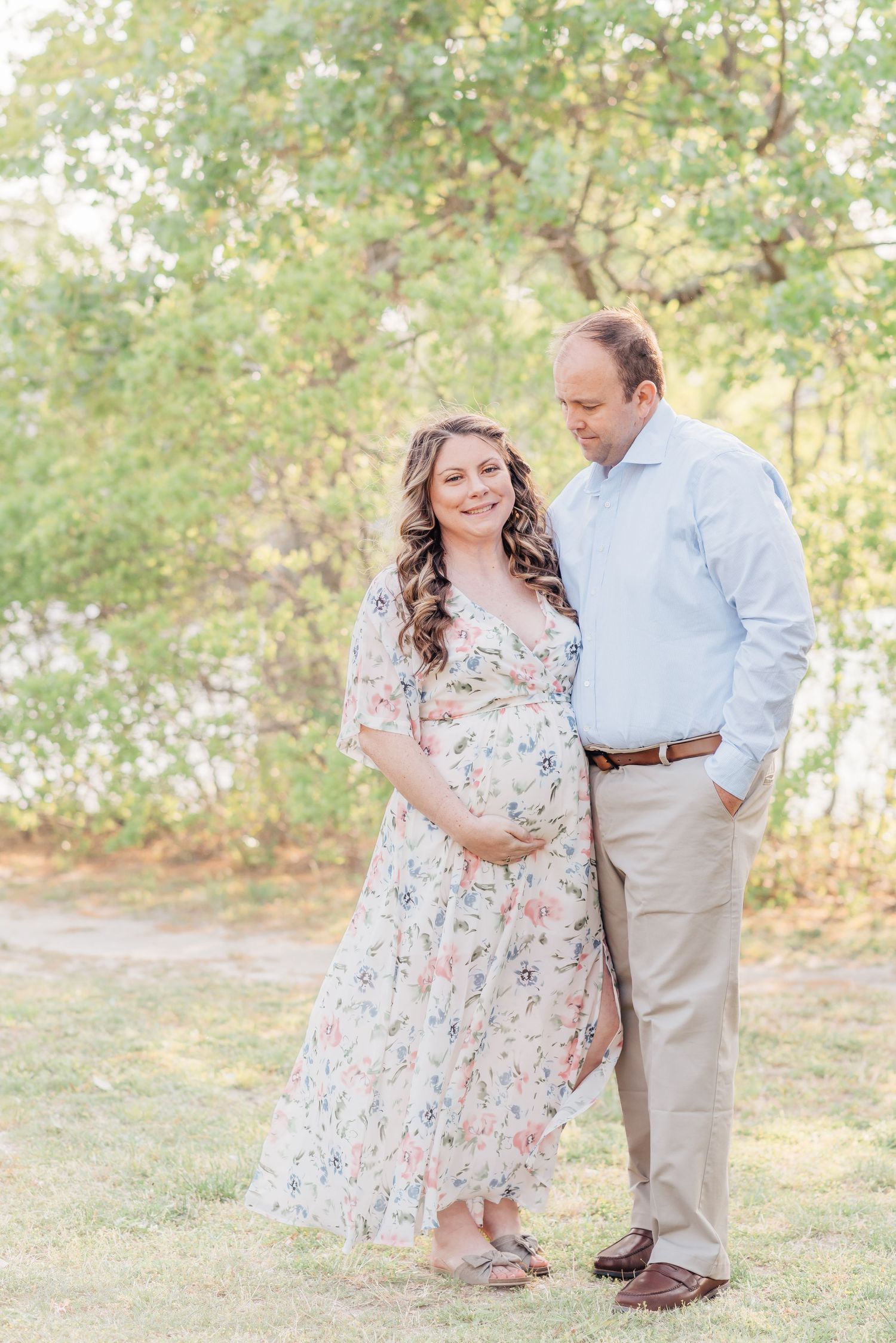 Portrait of a man and a pregnant woman outdoors leaning into each other looking down with her hands placed on her stomach and he is smiling.
