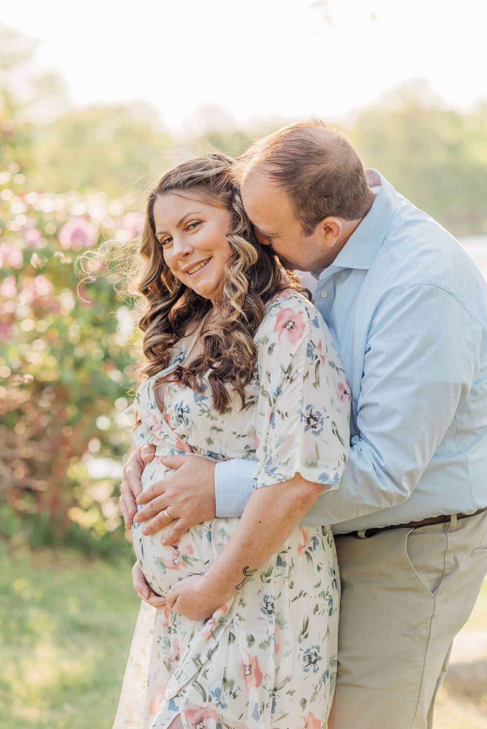 Portrait of a pregnant woman in a park wearing a floral dress, smiling while her husband wraps his arms around her.