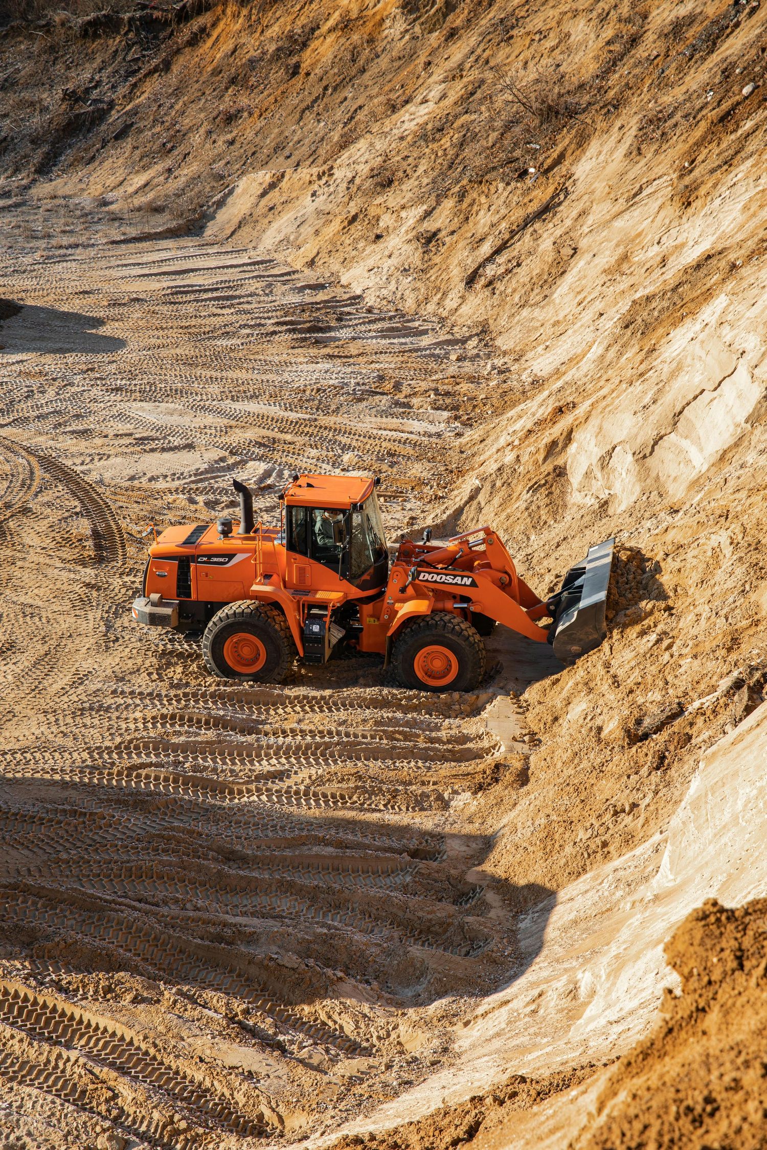 An orange front loader excavator operates in a sandy construction quarry with tire tracks visible in the soil.