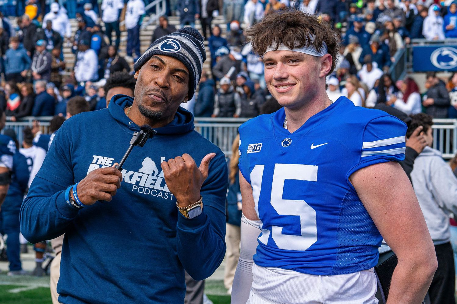 Two members of the Penn State football program interact on the field, one in a blue jersey numbered 15 and one in team apparel.