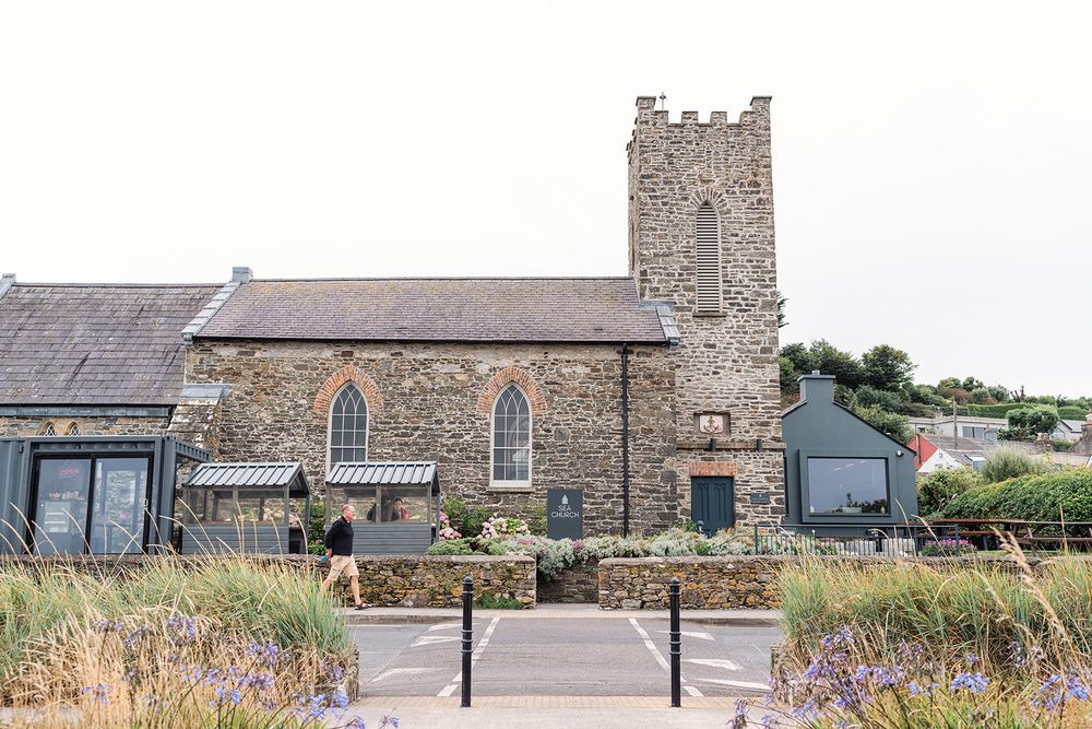 An exterior view of a stone church building with a tower and surrounding gardens.