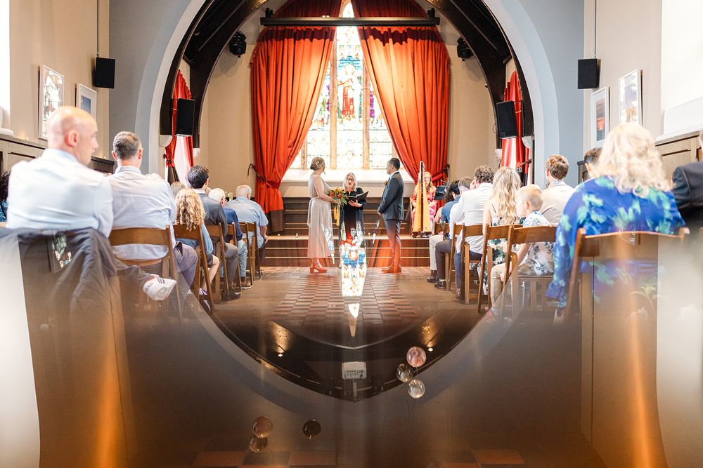 Wedding ceremony in progress viewed through arched doorway with orange curtains and checkered flooring.