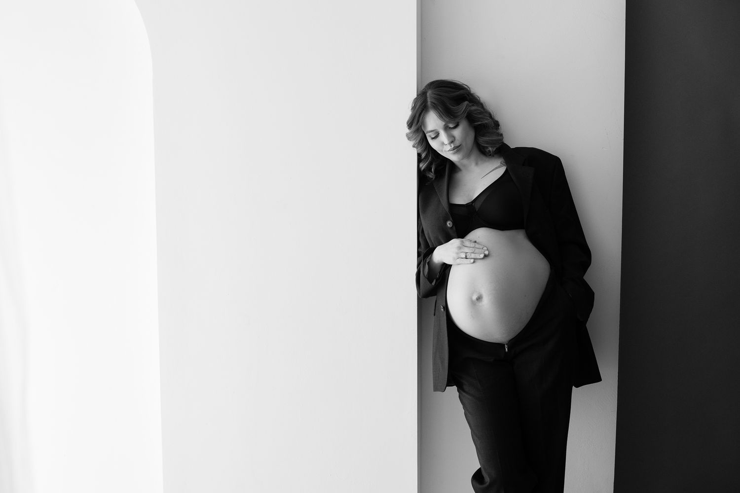 A black and white maternity photo showing an elegant side view of an expectant mother in dark clothing by a white wall.