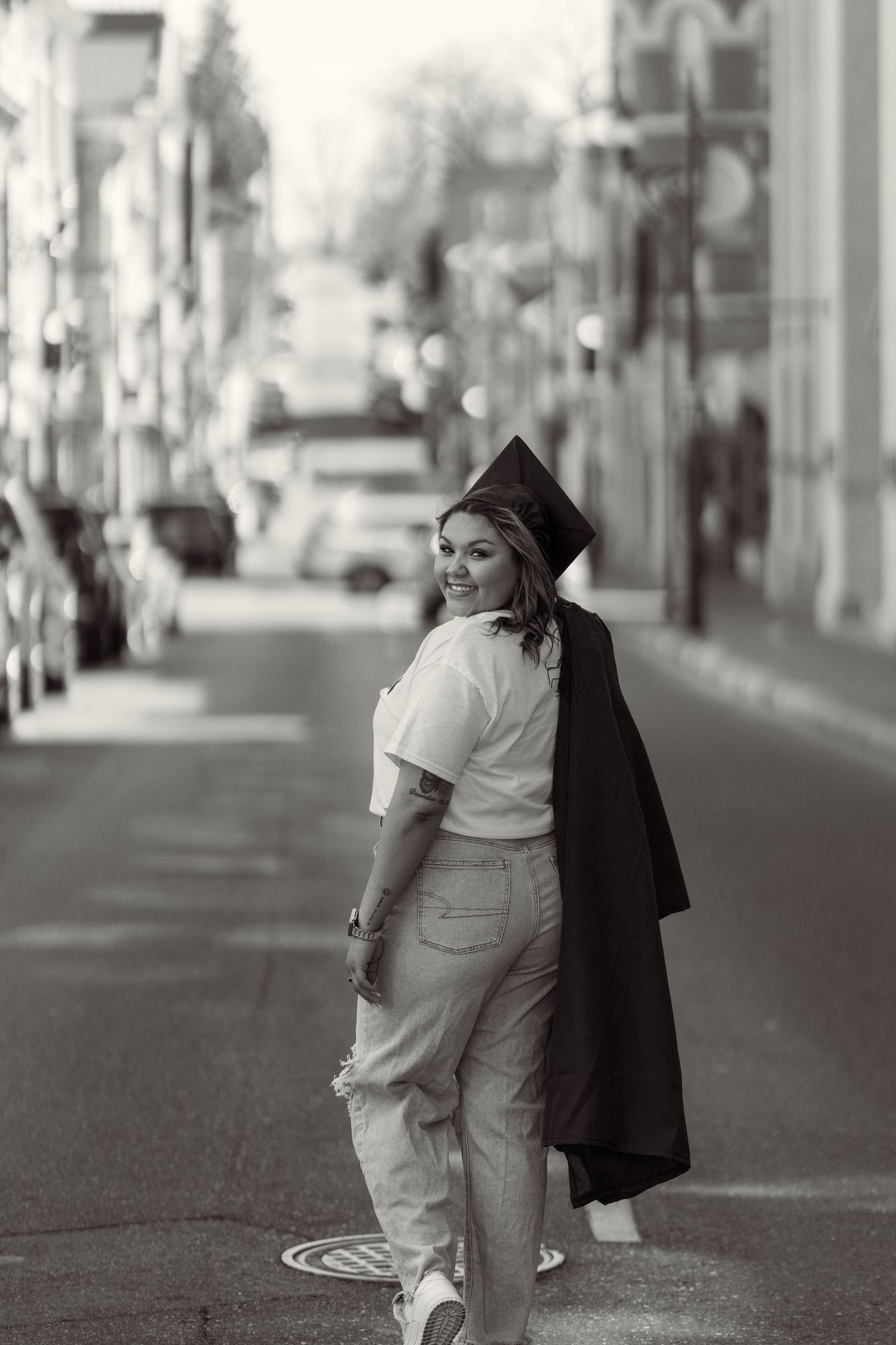 A graduate in a black cap and gown poses on a city street while looking over their shoulder in a black and white photo.
