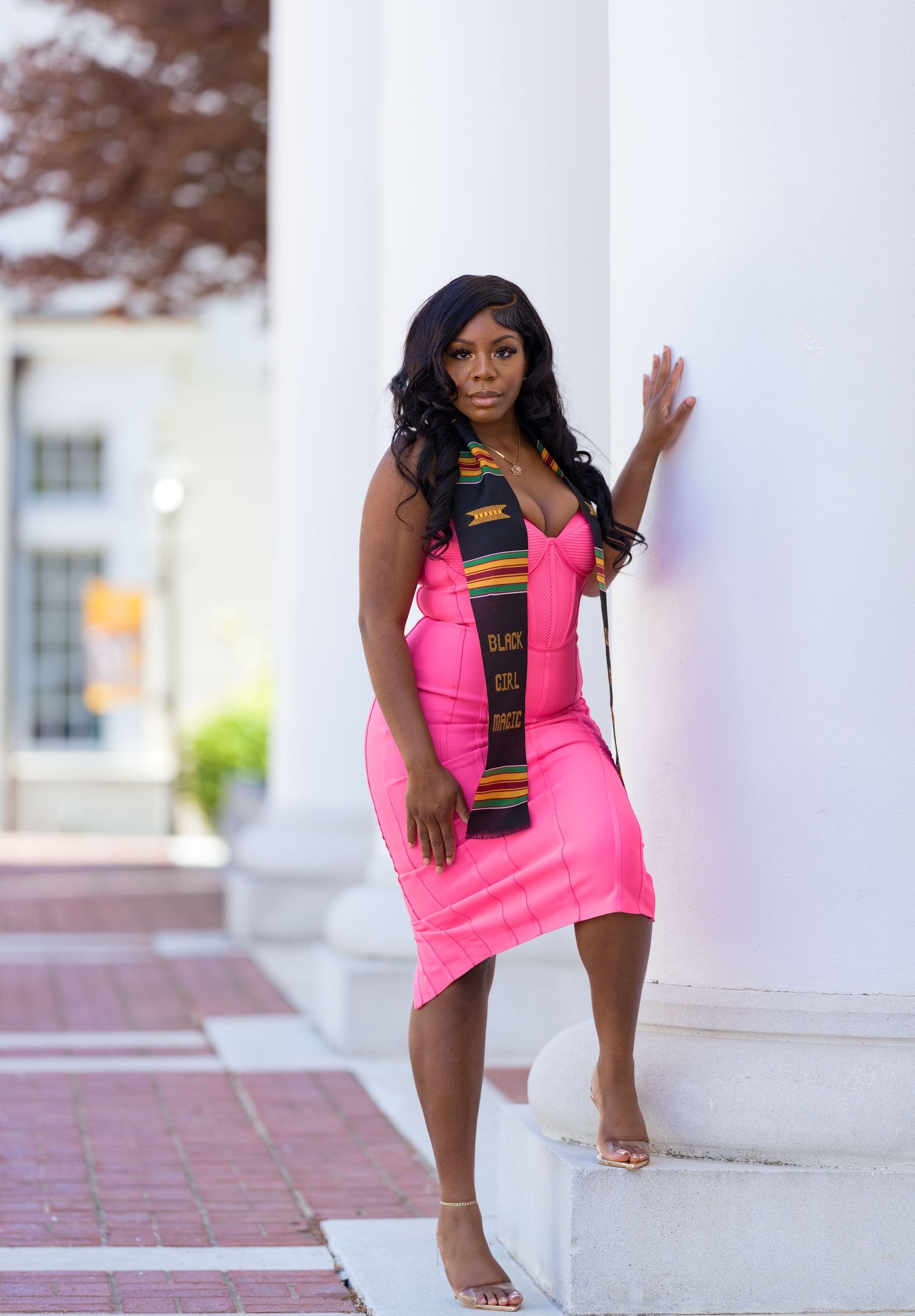 Woman in bright pink form-fitting dress with black and gold accent stripe poses against white wall outdoors.
