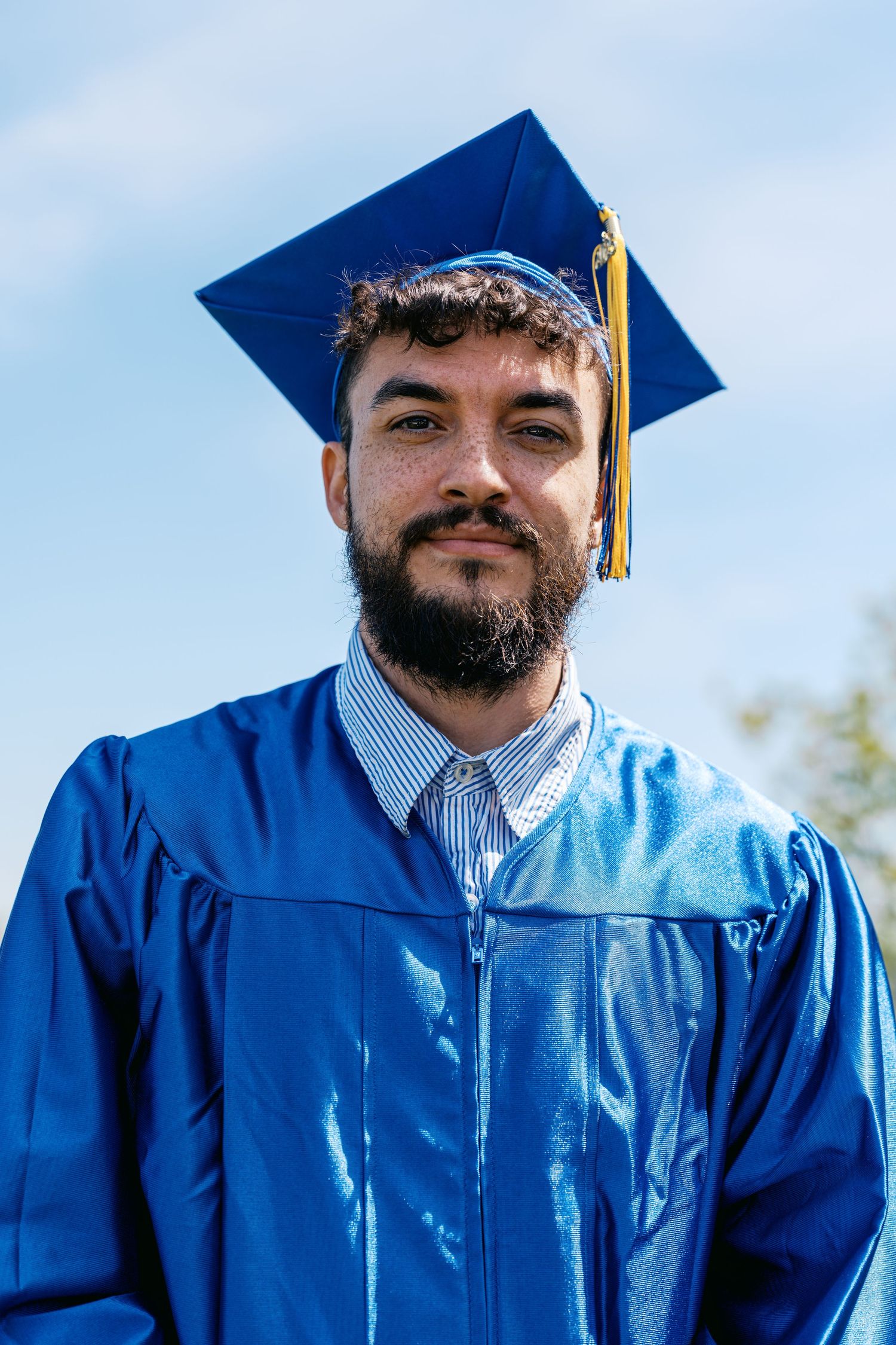 Graduate wearing bright blue cap and gown with tassel stands against sunny outdoor background.