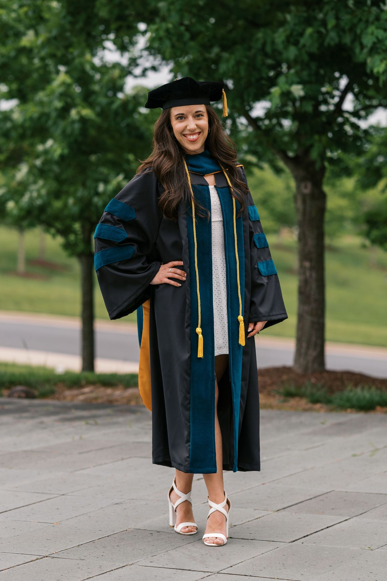 Graduate in black doctoral regalia with blue trim and gold sash poses outdoors in white heels on campus grounds.