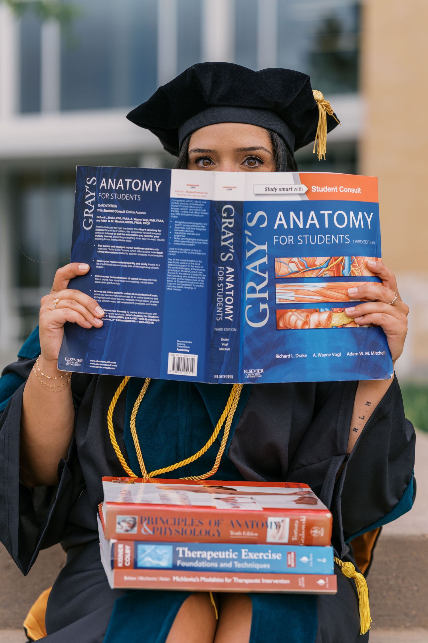 Graduate in cap and gown holds Gray's Anatomy textbooks and medical reference materials during graduation photo.
