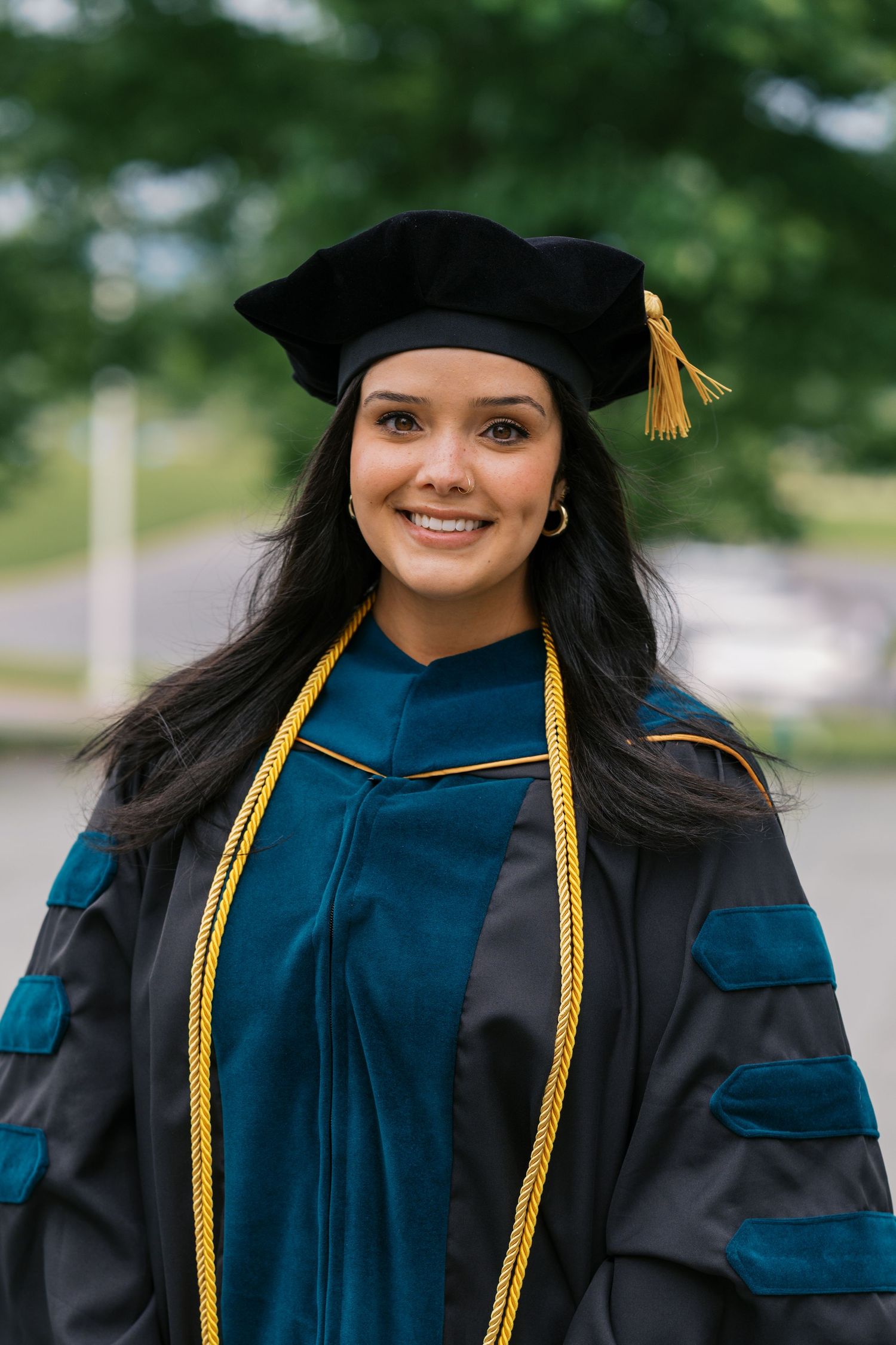Graduate wearing doctoral regalia with black cap, teal and grey gown, and gold honor cords smiling outdoors.