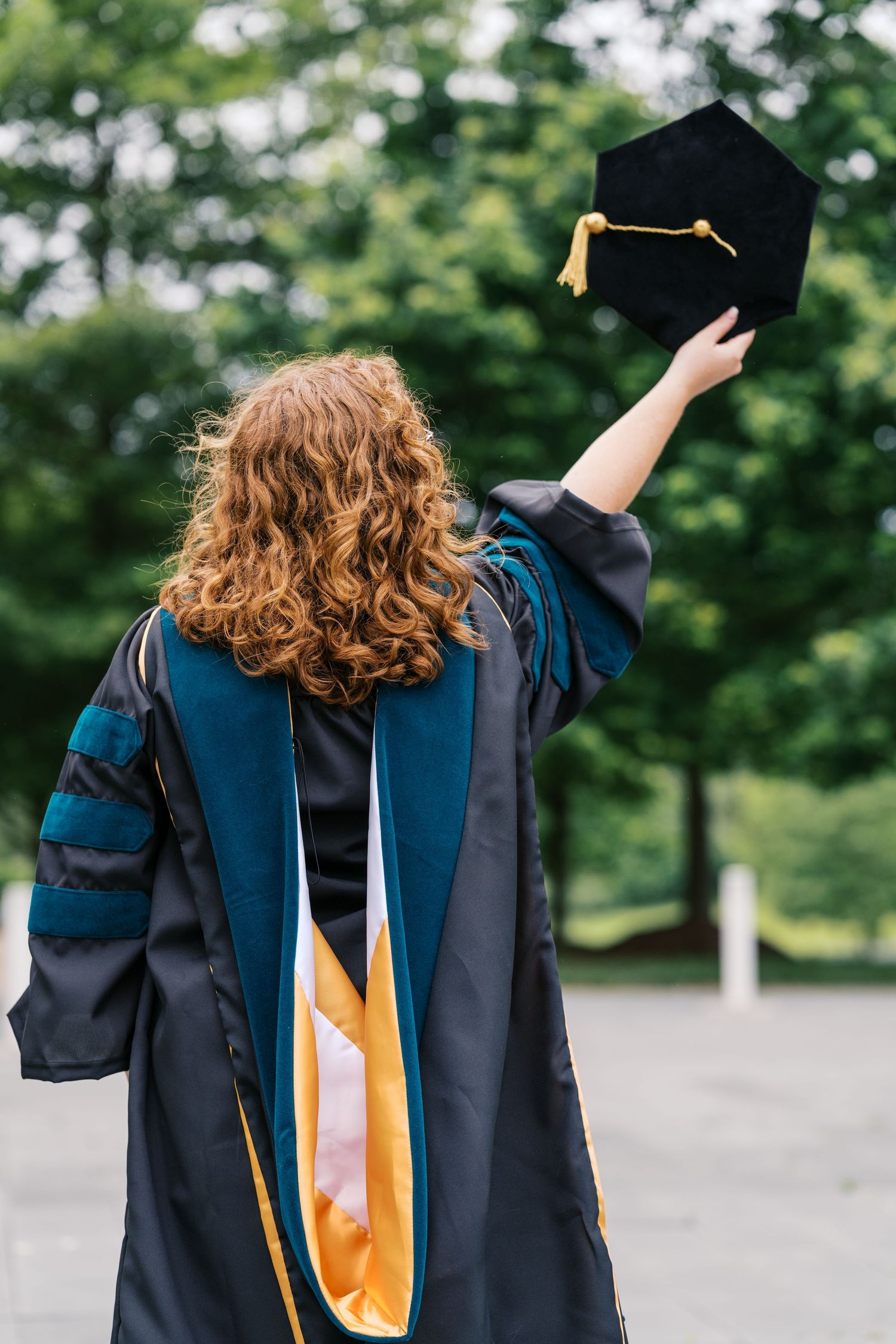 Graduate in blue and gold academic regalia holds up mortarboard cap against green foliage background.