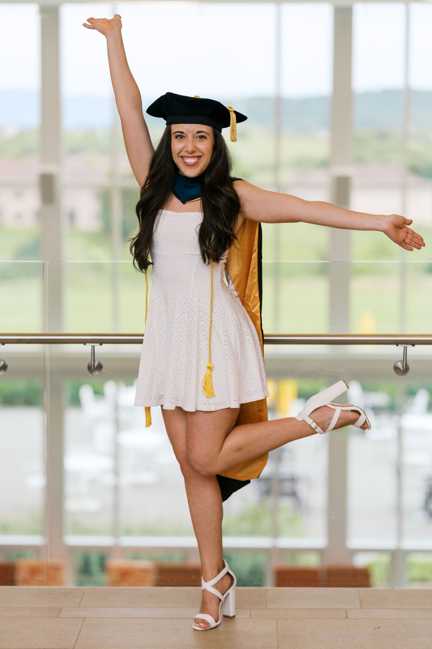 Graduate in white dress and cap strikes joyful pose with arms outstretched in bright indoor setting.