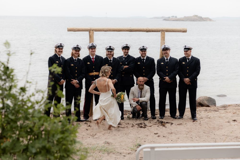 A wedding party poses together outdoors on a rocky coastal setting with groomsmen in dark uniforms against natural scenery.