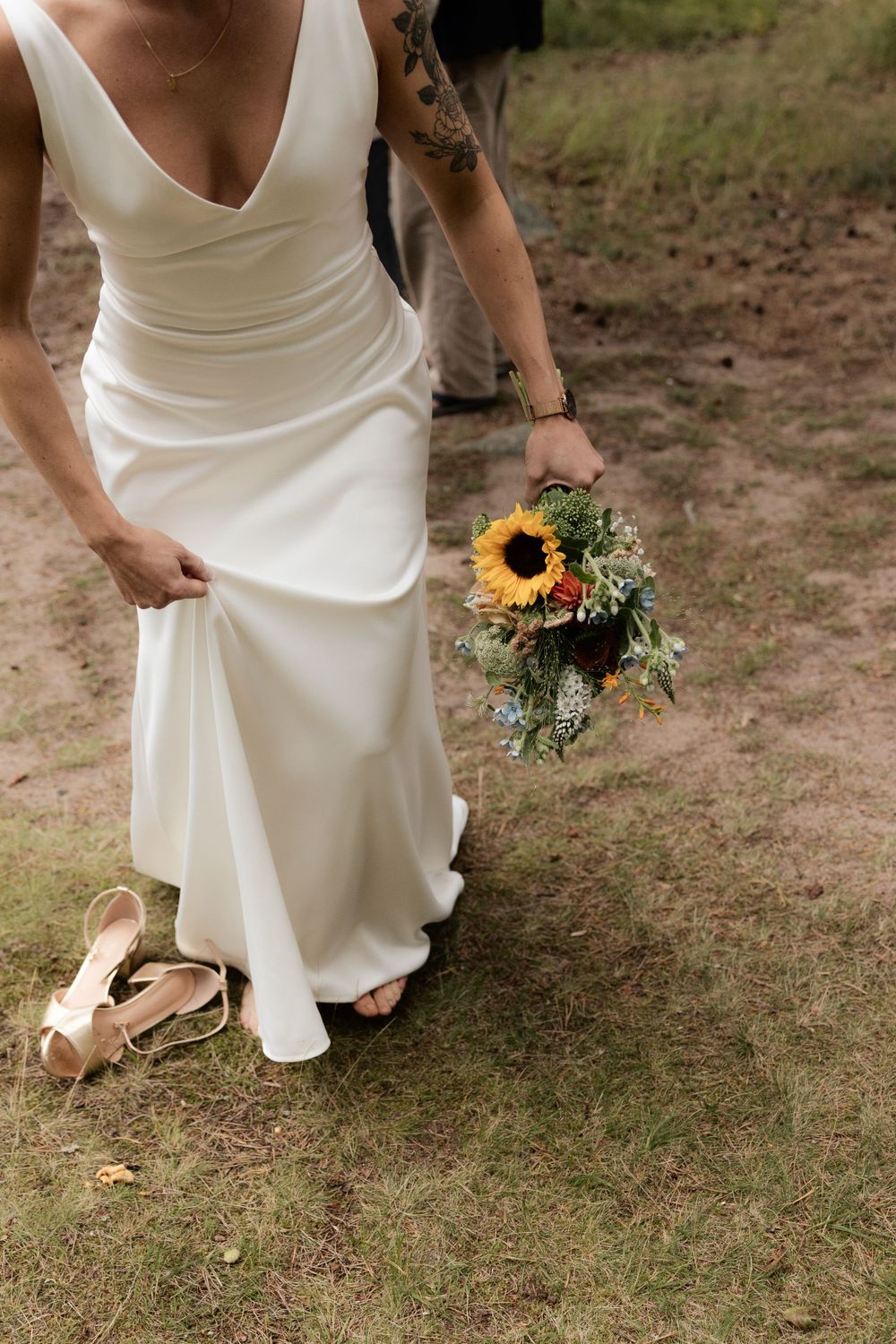 A white wedding dress and sunflower bouquet against rustic outdoor setting with sandals on grass.