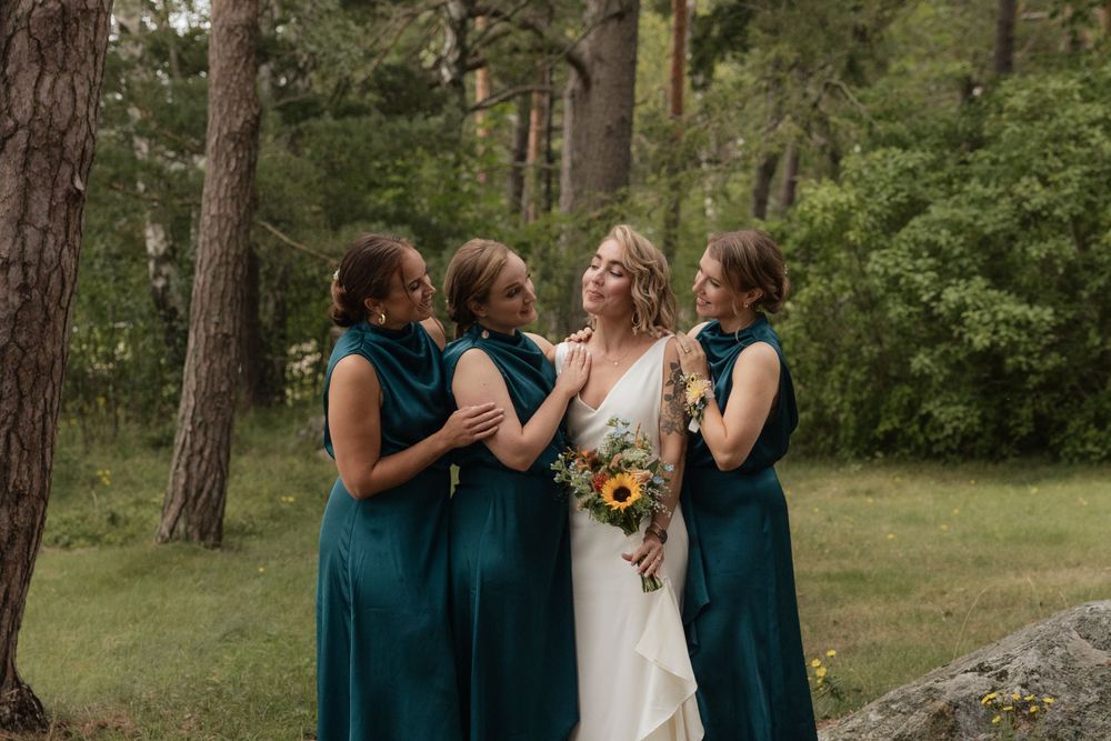 A wedding party in teal bridesmaid dresses and a white bridal gown share a joyful moment in a wooded outdoor setting.
