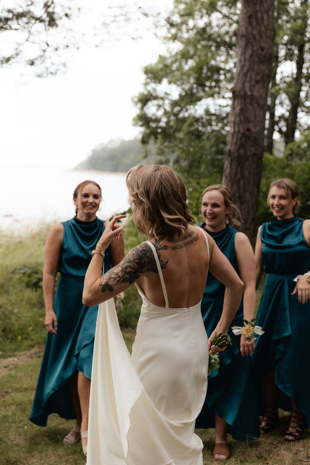 Bridesmaids in teal dresses share a joyful moment with bride in white backless gown during outdoor forest wedding.