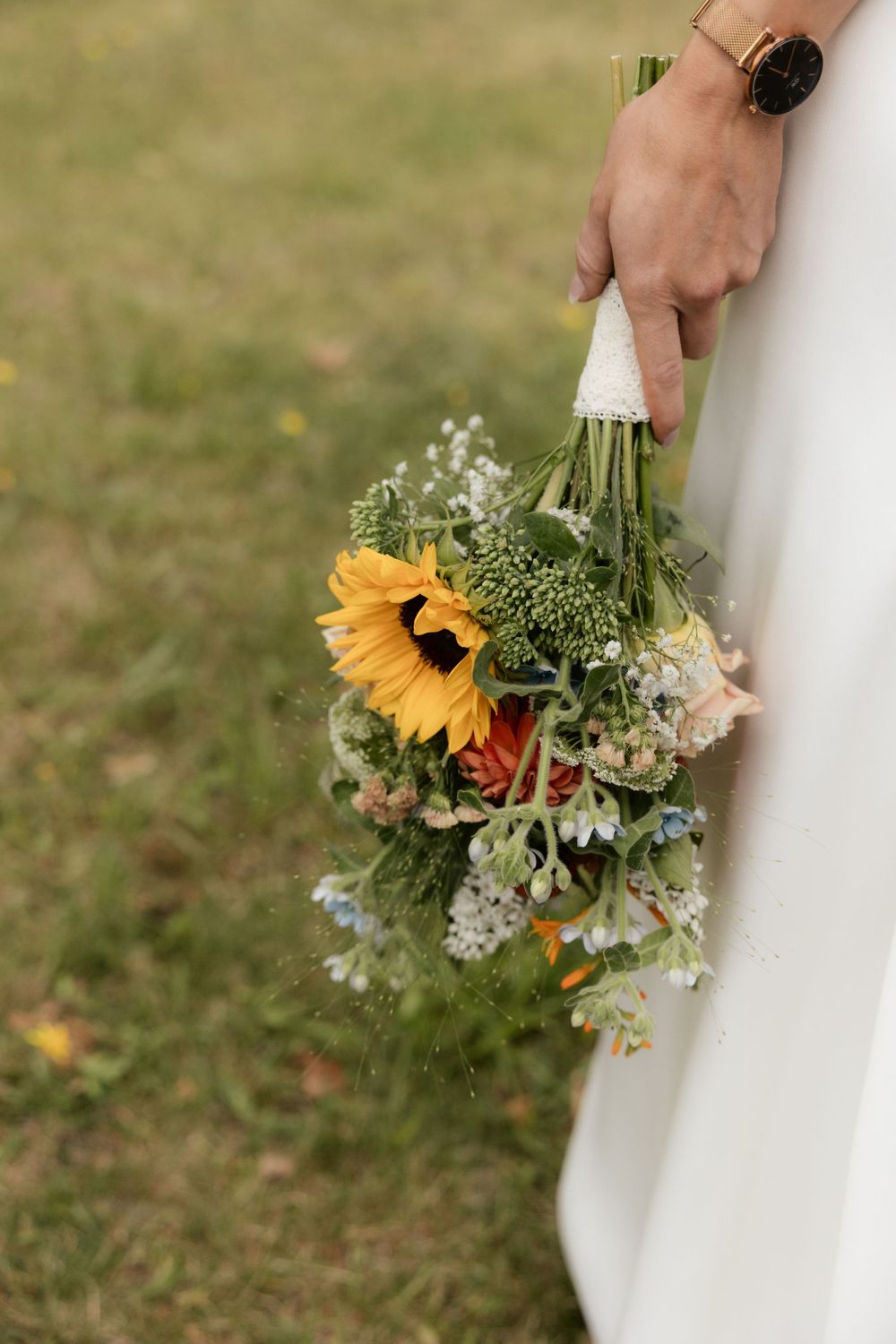 Hand holding rustic bouquet with bright sunflower, white florals and greenery against grassy background.