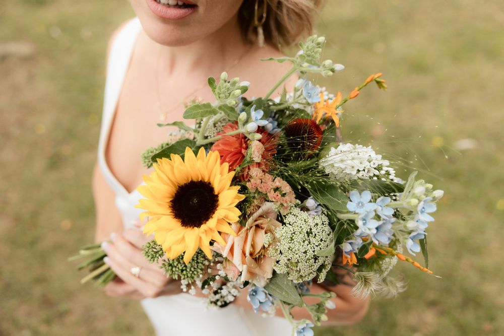 A summery wildflower bouquet featuring a bright sunflower, delicate blue blooms, and peach roses against a blurred background.