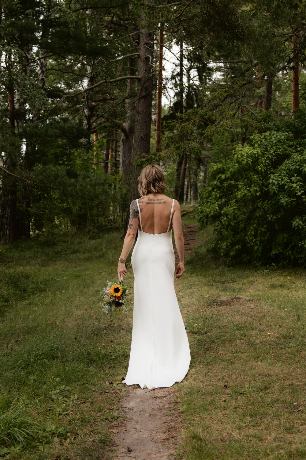 A bride in a backless white wedding dress holds a sunflower bouquet while walking on a forest trail.