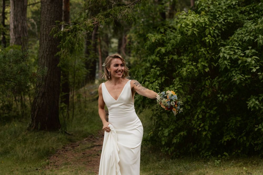 A bride in a simple white wedding dress holds a bouquet while standing on a wooded path.