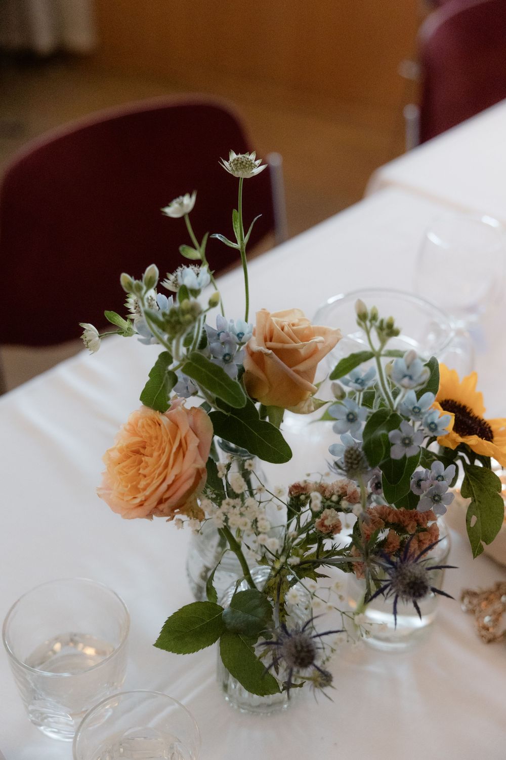 Elegant peach roses and delicate blue flowers arranged in clear glass vases on a white tablecloth with candlelight.