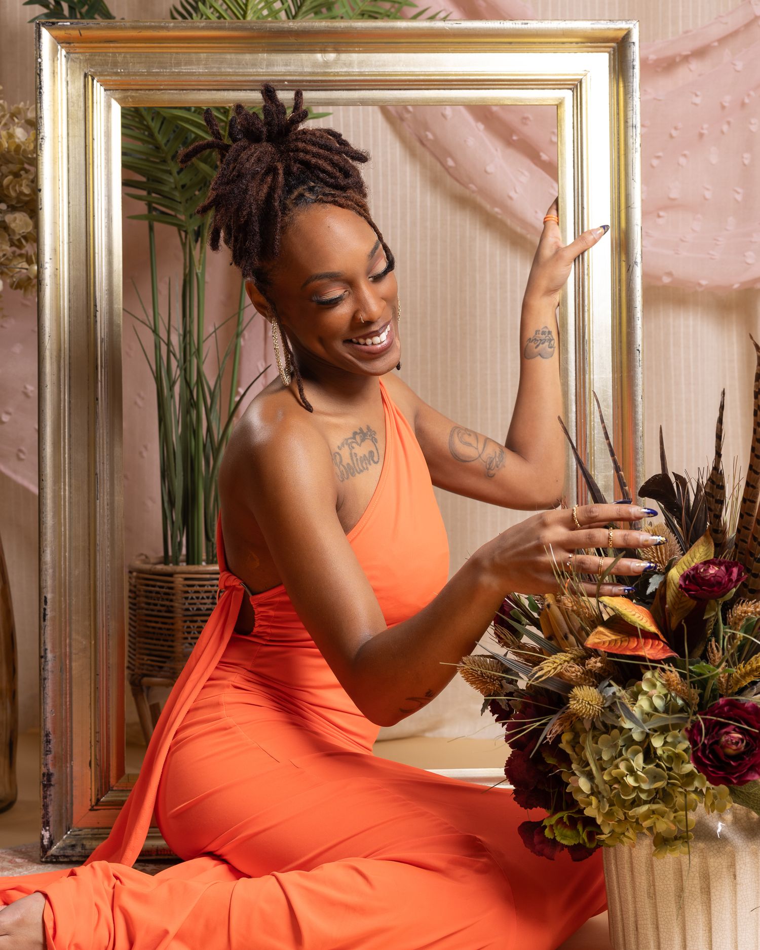 Person in vibrant orange dress laughs joyfully while sitting next to plants and decorative frame in warm interior setting.