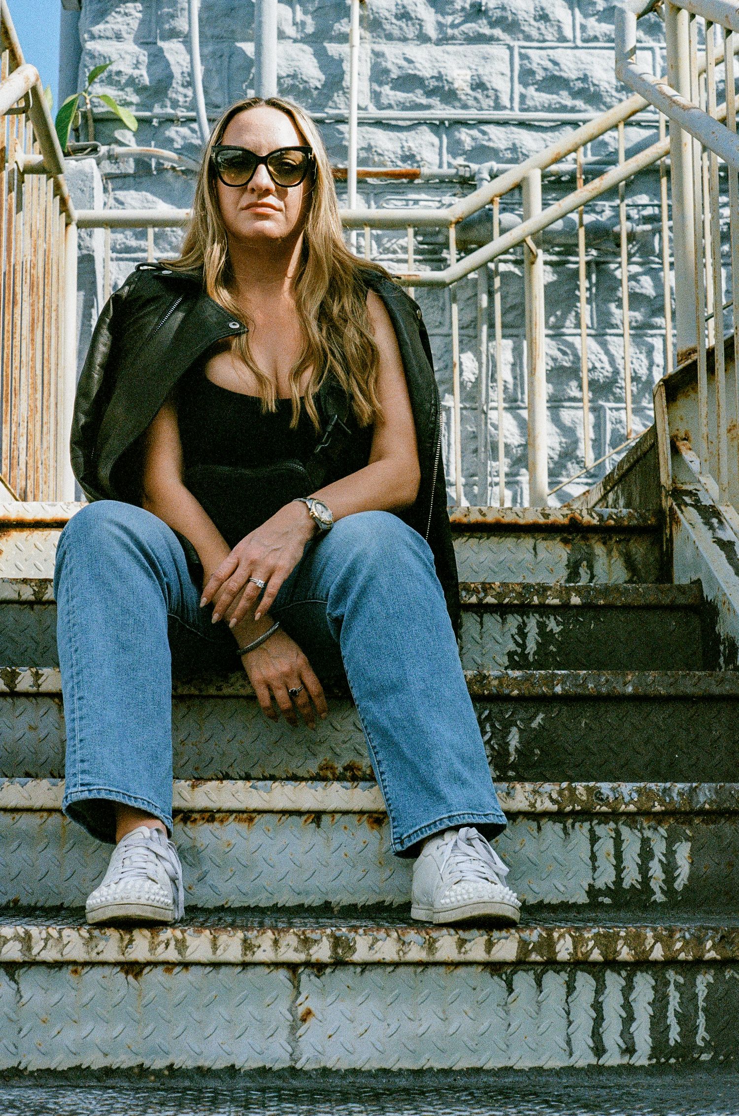 Fashion portrait of someone sitting on concrete steps wearing sunglasses, a black top and jeans against stone wall backdrop.