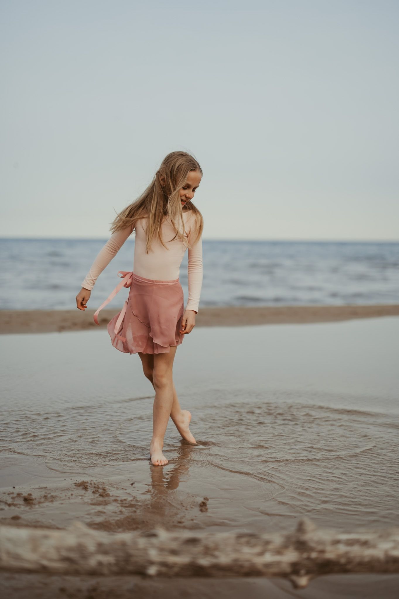 Young girl in pink skirt and white top walks barefoot along a sandy beach shoreline with calm ocean waves.