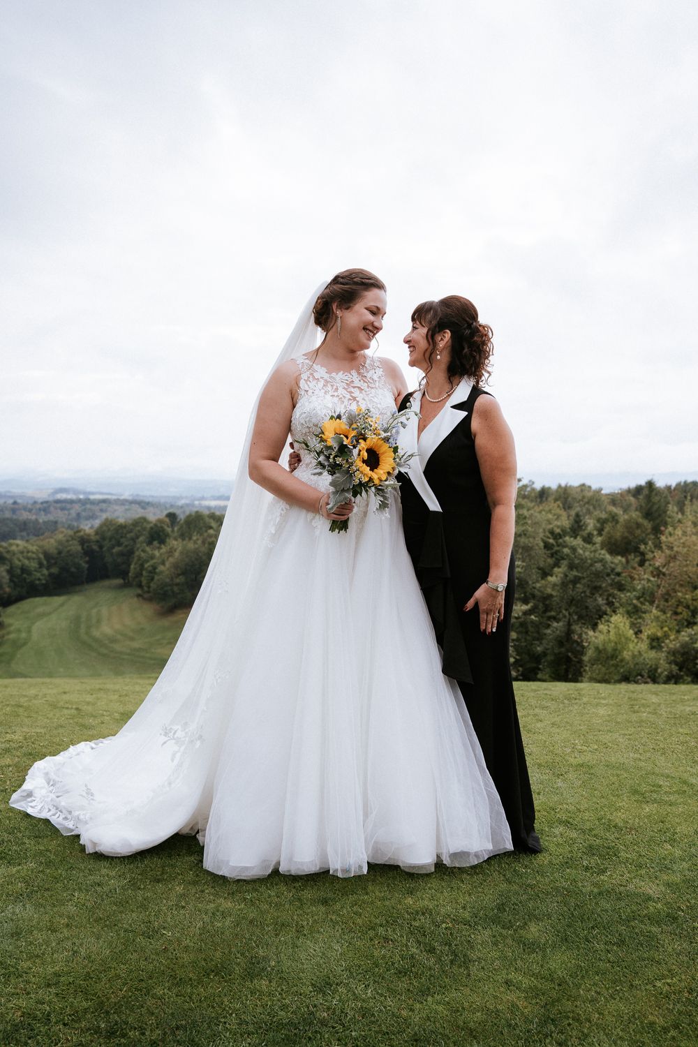 Deux personnes partagent un moment heureux au sommet d'une colline en tenue de mariage, entourées de tournesols sous un ciel nuageux.