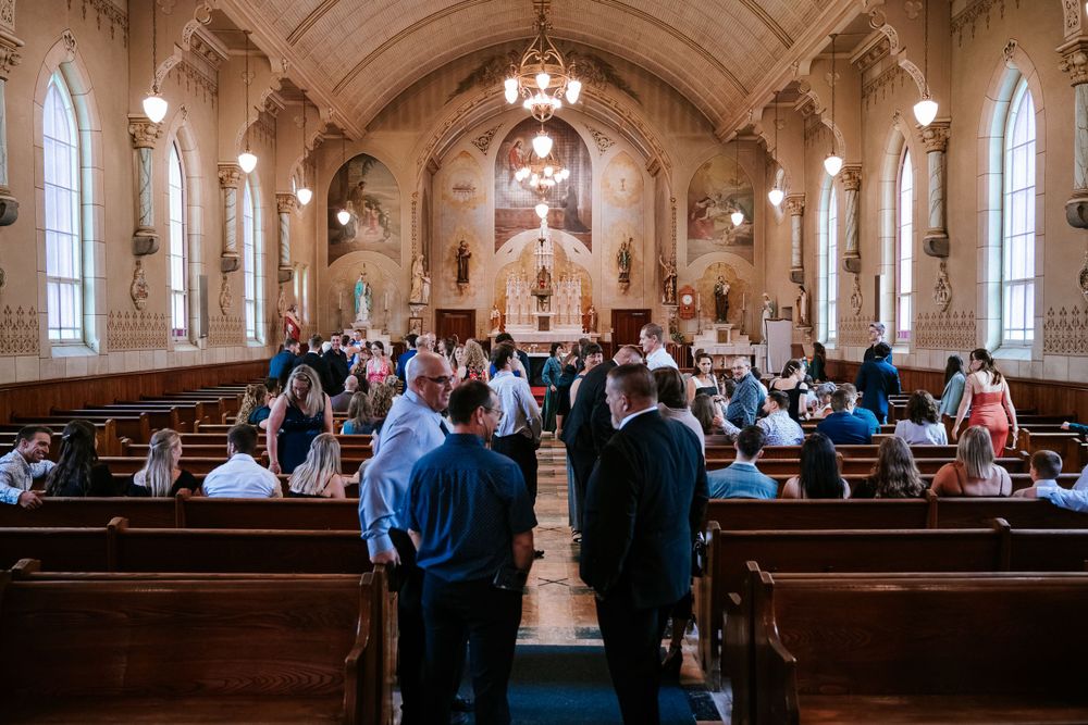 Des invités de mariage remplissent les bancs en bois d'une grande cathédrale, avec des plafonds voûtés, des vitraux et des lustres élégants.