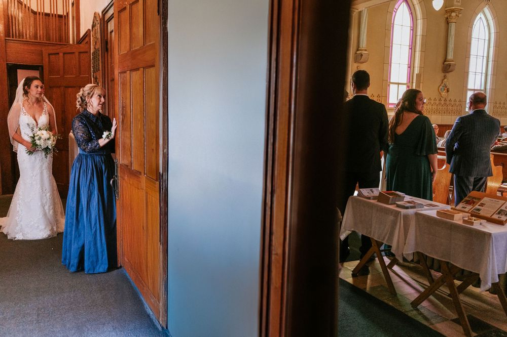 Mariée en robe blanche avec une demoiselle d’honneur en robe bleue, debout devant les portes en bois de l’église avant la cérémonie.