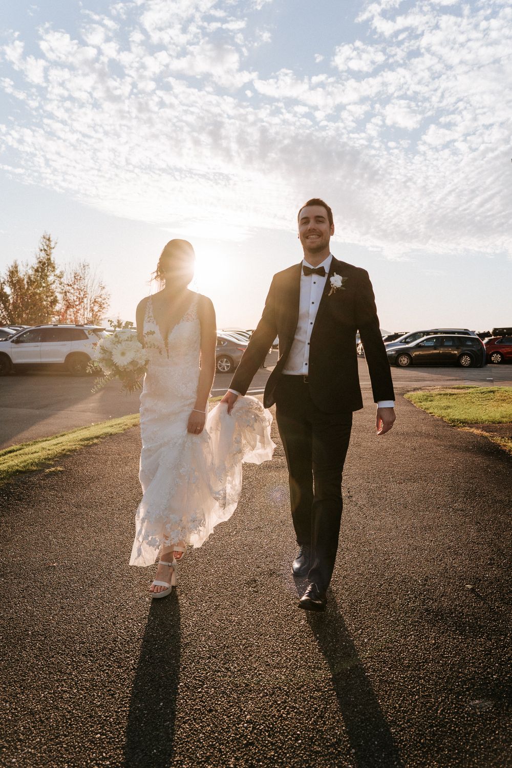Les mariés marchent main dans la main sur un chemin baigné de soleil, la mariée en robe de dentelle tenant son bouquet, le marié en smoking.