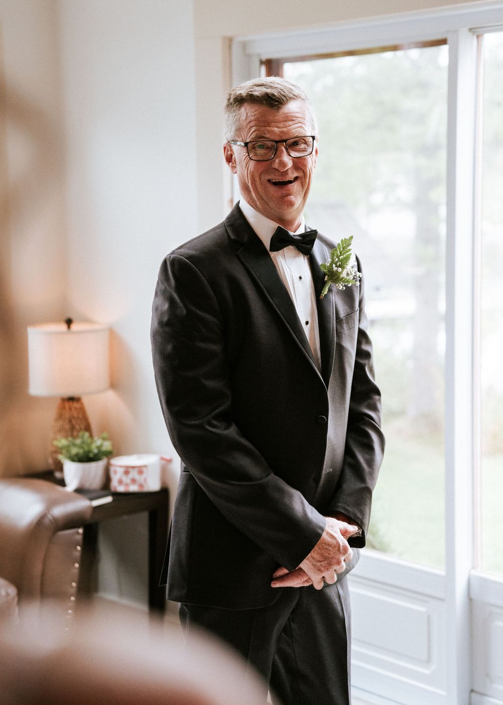Une personne en smoking noir classique avec nœud papillon et boutonnière pose près d'une fenêtre dans un cadre formel.