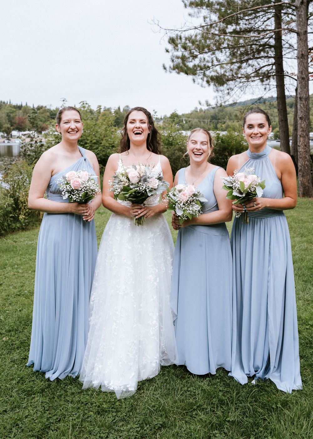 Un groupe de mariage en robes bleu poudre et robe blanche tient des bouquets, posant sur l'herbe près des pins.