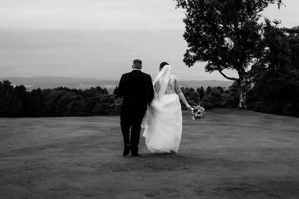 Un couple en tenue de mariage marche sur une colline avec vue sur un paysage montagneux, en noir et blanc.