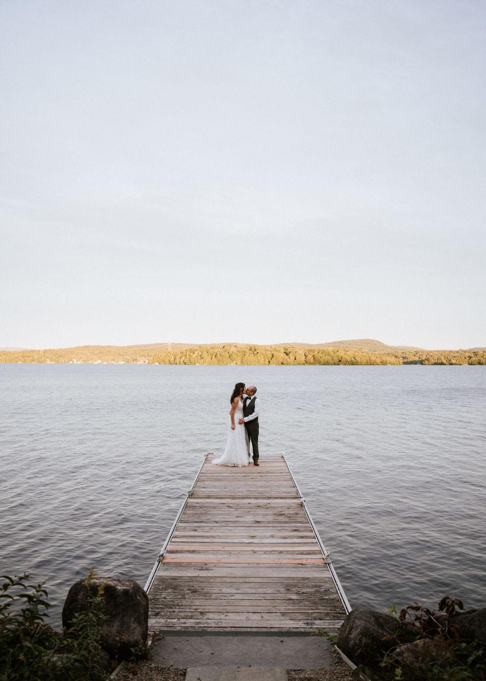Couple sur ponton en bois, vue sur lac serein et collines au loin.